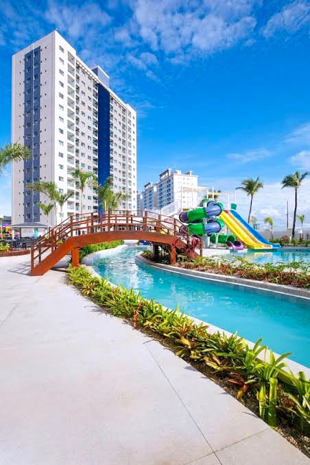 A vibrant resort pool area is seen with lush greenery lining the walkway. A wooden bridge crosses over a winding lazy river. In the distance, a waterslide adds a playful element to the space, complemented by tall palm trees against a clear blue sky.