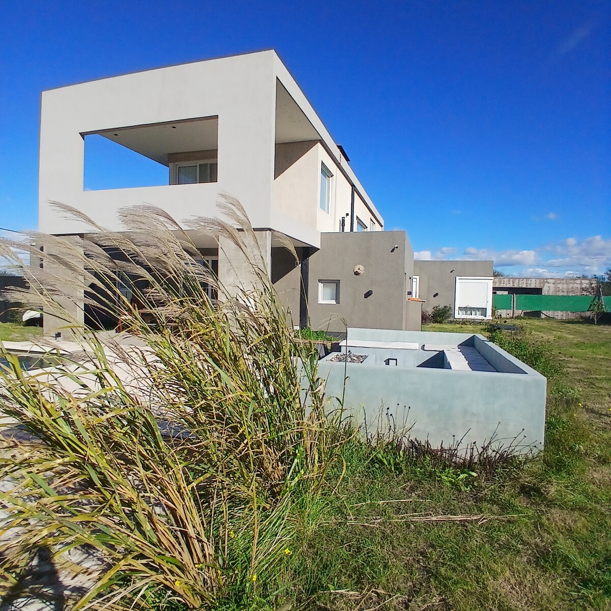 A modern two-story building is surrounded by a natural landscape, featuring tall grass in the foreground. A light gray structure with a rectangular design is visible, complemented by large windows. The open area offers views of clear blue skies and distant greenery.