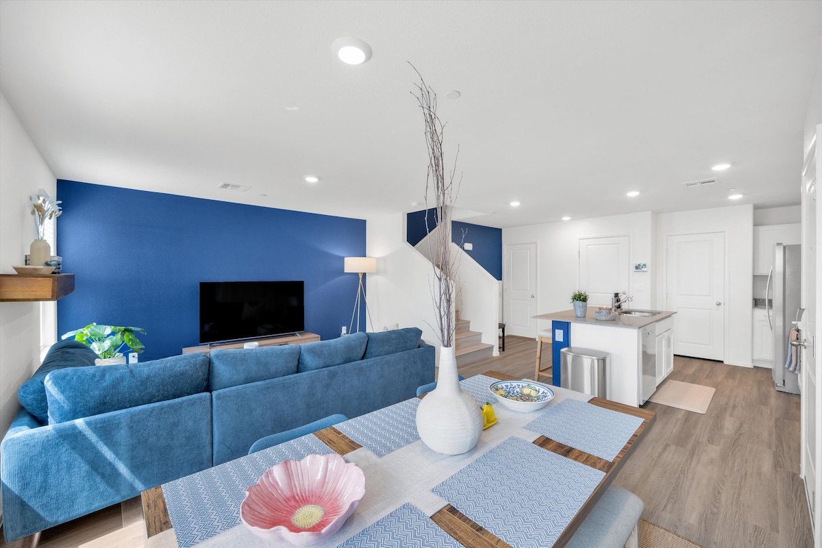 An expansive open-concept living area showcases a comfortable blue sectional sofa facing a large television. A dining table with a vibrant floral centerpiece is featured in the foreground, accompanied by modern kitchen appliances visible in the background. Light-colored walls contrast with deep blue accent walls.
