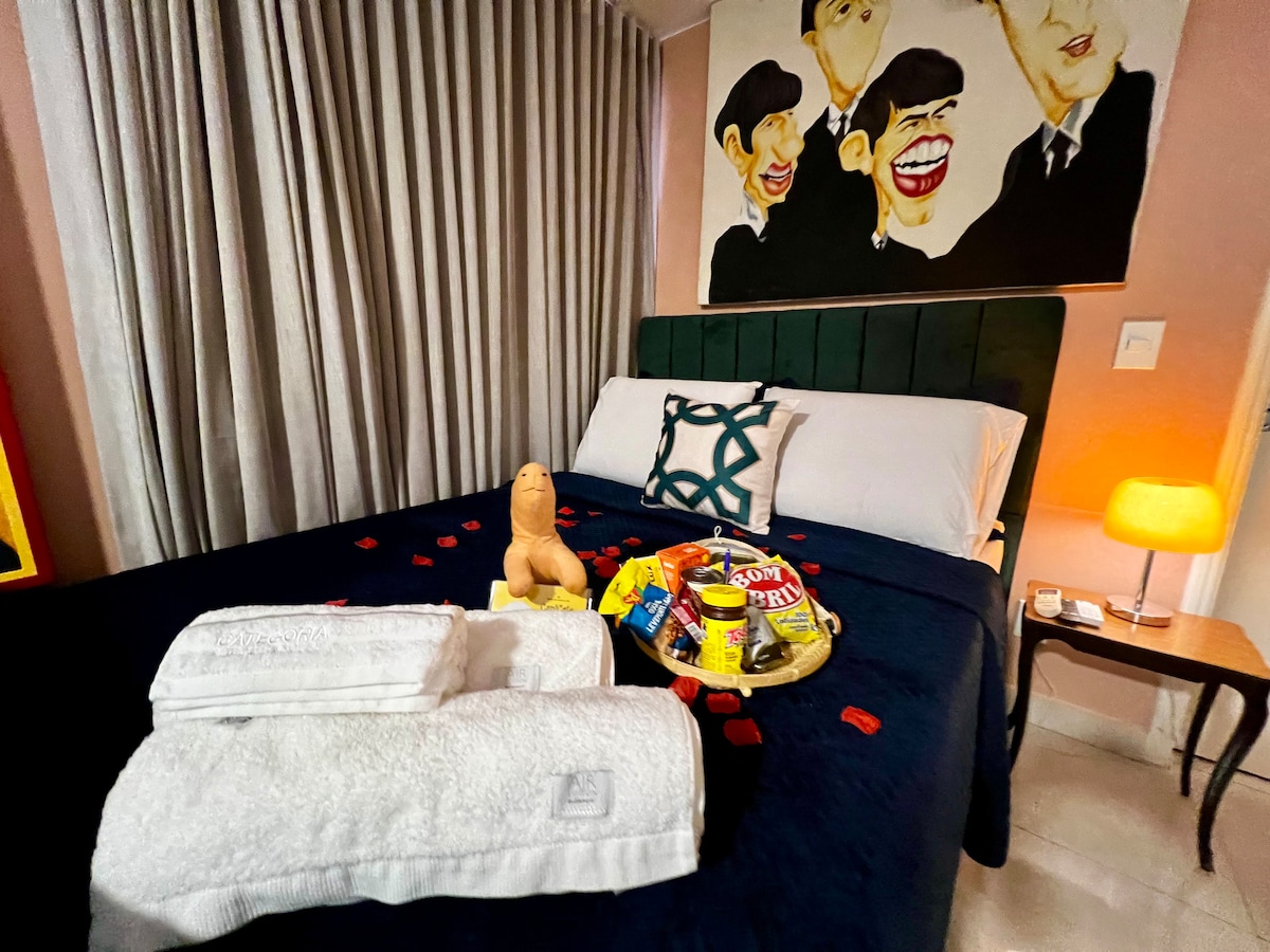 A spacious bed with a dark blue bedspread is adorned with towels and a tray filled with snacks. An artwork featuring iconic figures hangs above the bed, while soft light from a lamp illuminates the corner. Drapes frame the window, enhancing the cozy atmosphere.