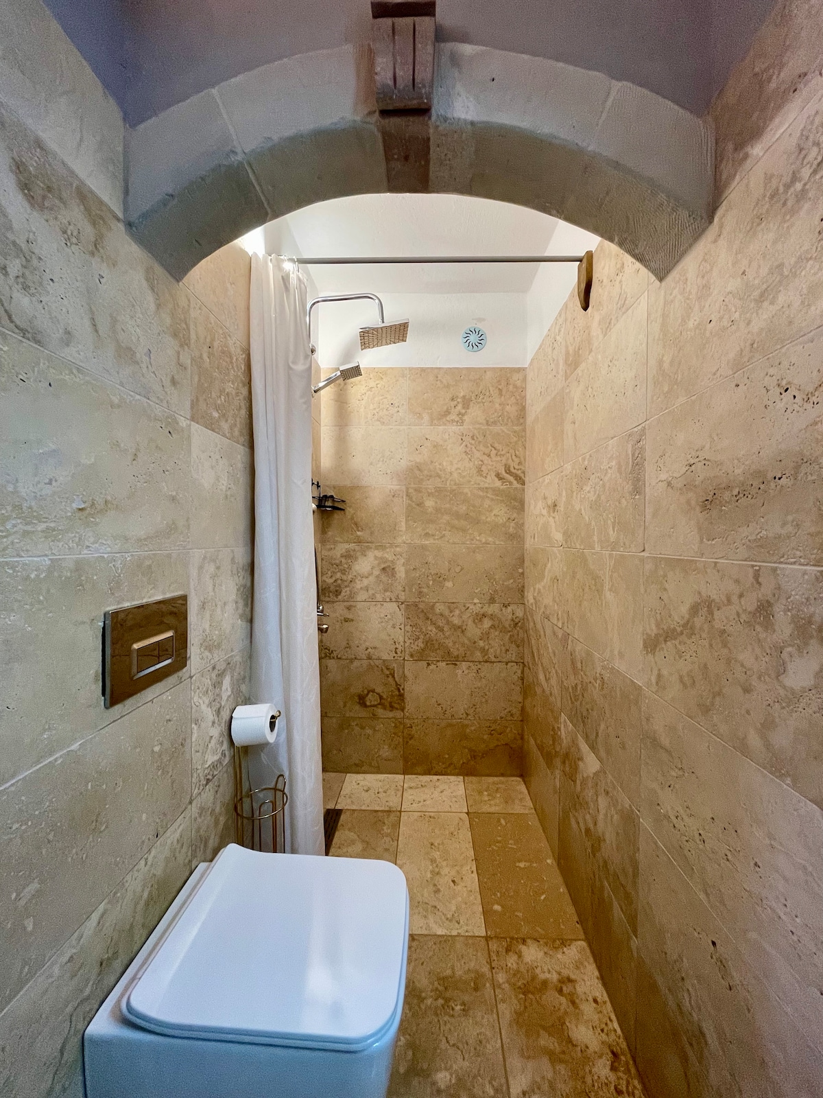 A sleek bathroom is highlighted by natural stone wall tiles. A spacious shower is equipped with a modern shower head and a white curtain. A compact toilet is positioned near the wall, and a roll of toilet paper is provided for convenience.
