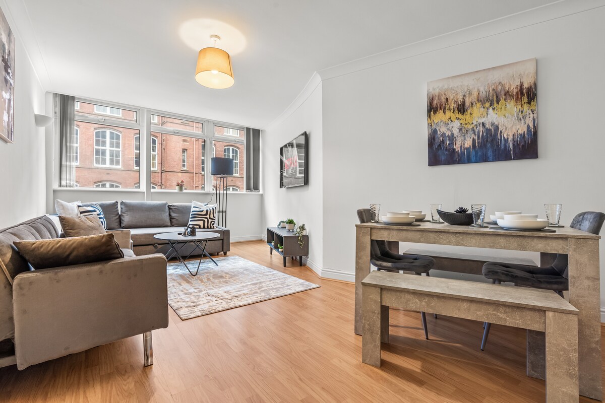 A spacious lounge area features contemporary furnishings, including a comfortable sofa and a sleek coffee table. A dining table with seating for six is positioned nearby. Large windows allow natural light to fill the room, highlighting a piece of modern wall art.