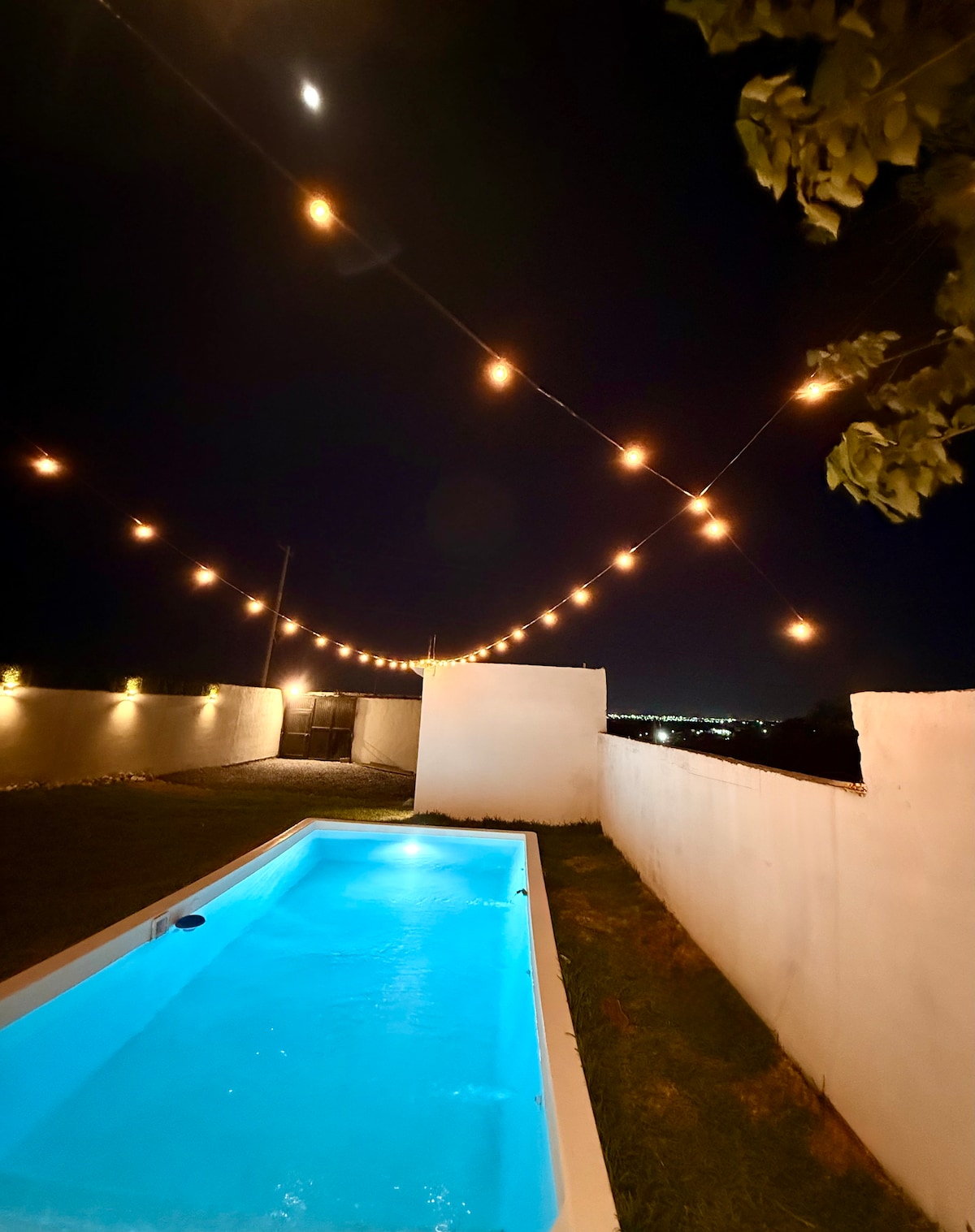 An inviting outdoor space features a pool with illuminated water reflecting a soft blue hue. String lights overhead create a warm ambiance against a night sky, enhancing the atmosphere around a well-kept grass area and white wall boundaries.