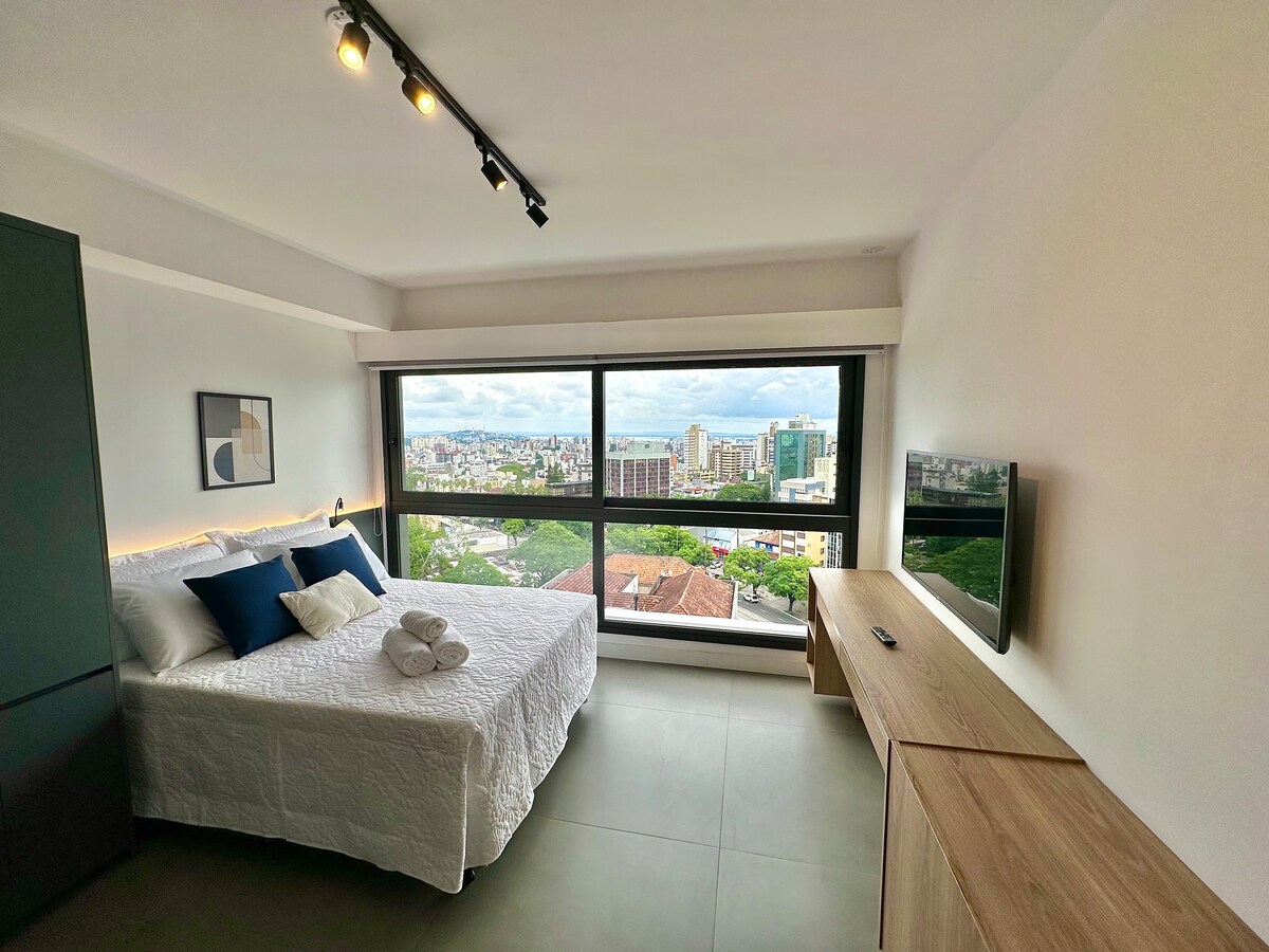 A bright and spacious sleeping area features a large bed adorned with white bedding and coordinating blue accents. A view of the city can be seen through expansive windows, while a wooden entertainment unit and wall-mounted TV add to the functionality of the space.