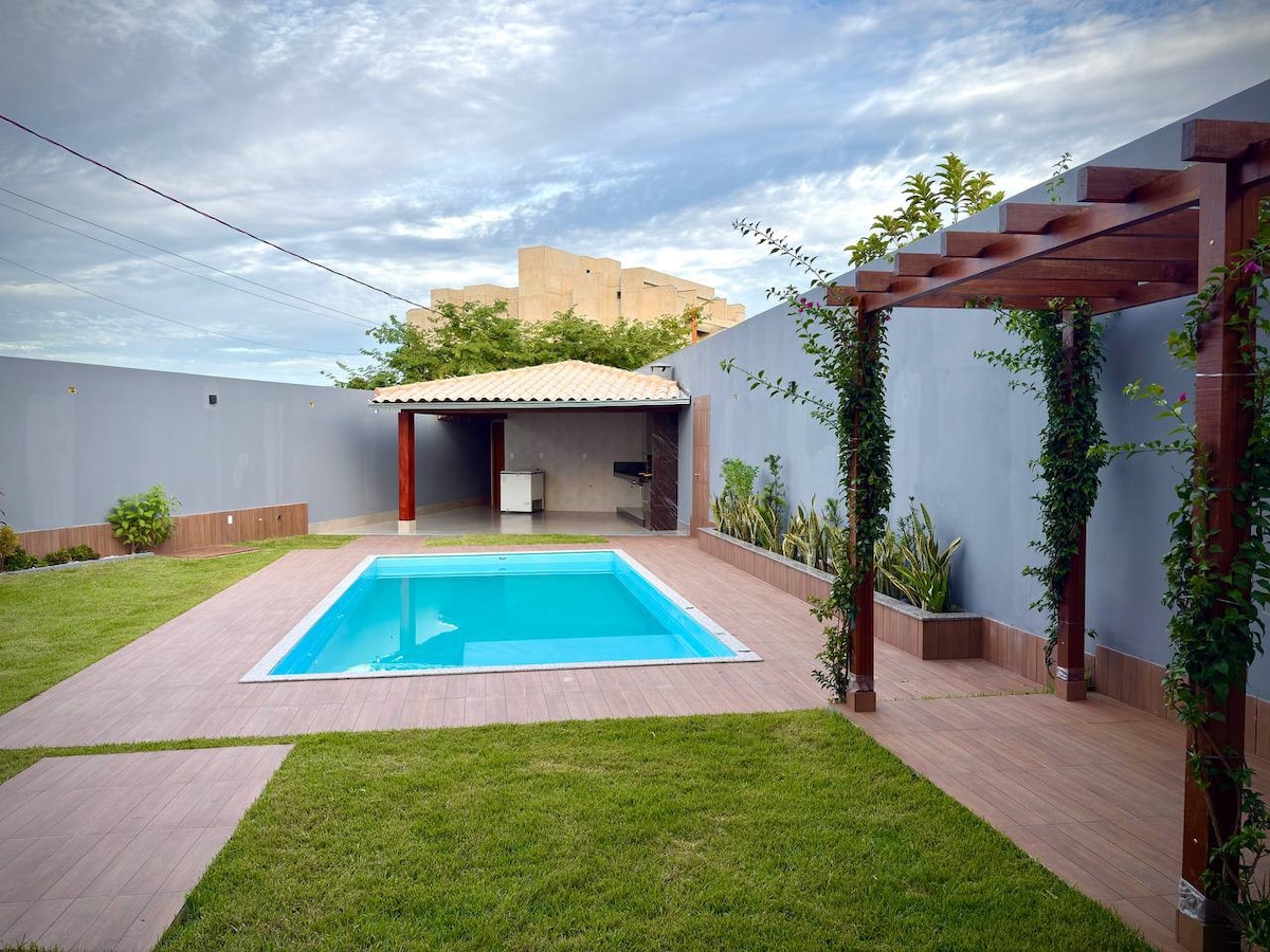 An outdoor area features a sparkling pool surrounded by wooden decking and neatly trimmed grass. Lush greenery and potted plants frame the pool, and a shaded pergola with climbing vines is visible, providing a relaxing space for outdoor gatherings. A gazebo is seen in the background.
