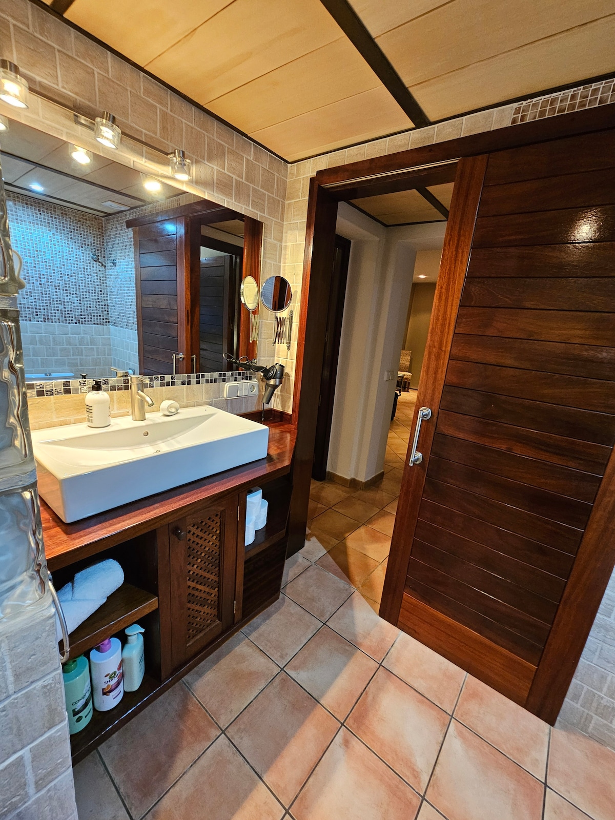A spacious bathroom is designed with natural stone tiles and warm wooden accents. A large sink with a modern faucet is placed on a wooden countertop, complemented by a wall mirror. Shelves are stocked with toiletries, and a door leads to an adjoining area.