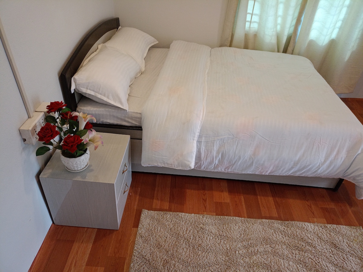 A neatly arranged sleeping area is featured, showcasing a spacious bed layered with white linens and two pillows. A light-colored bedside table sits beside the bed, adorned with a decorative planters holding artificial flowers. Soft natural light filters through the sheer curtains.