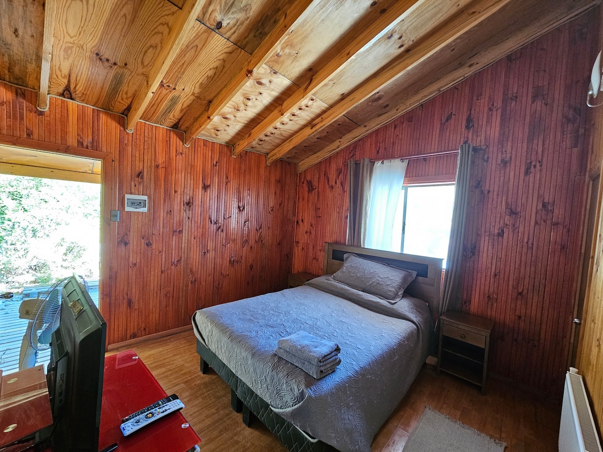 A welcoming bedroom features wooden paneling and exposed beams on the ceiling. A spacious bed is dressed in light gray bedding, complemented by neatly folded towels on top. A small bedside table stands next to the bed, with a television positioned across the room, offering a cozy retreat.