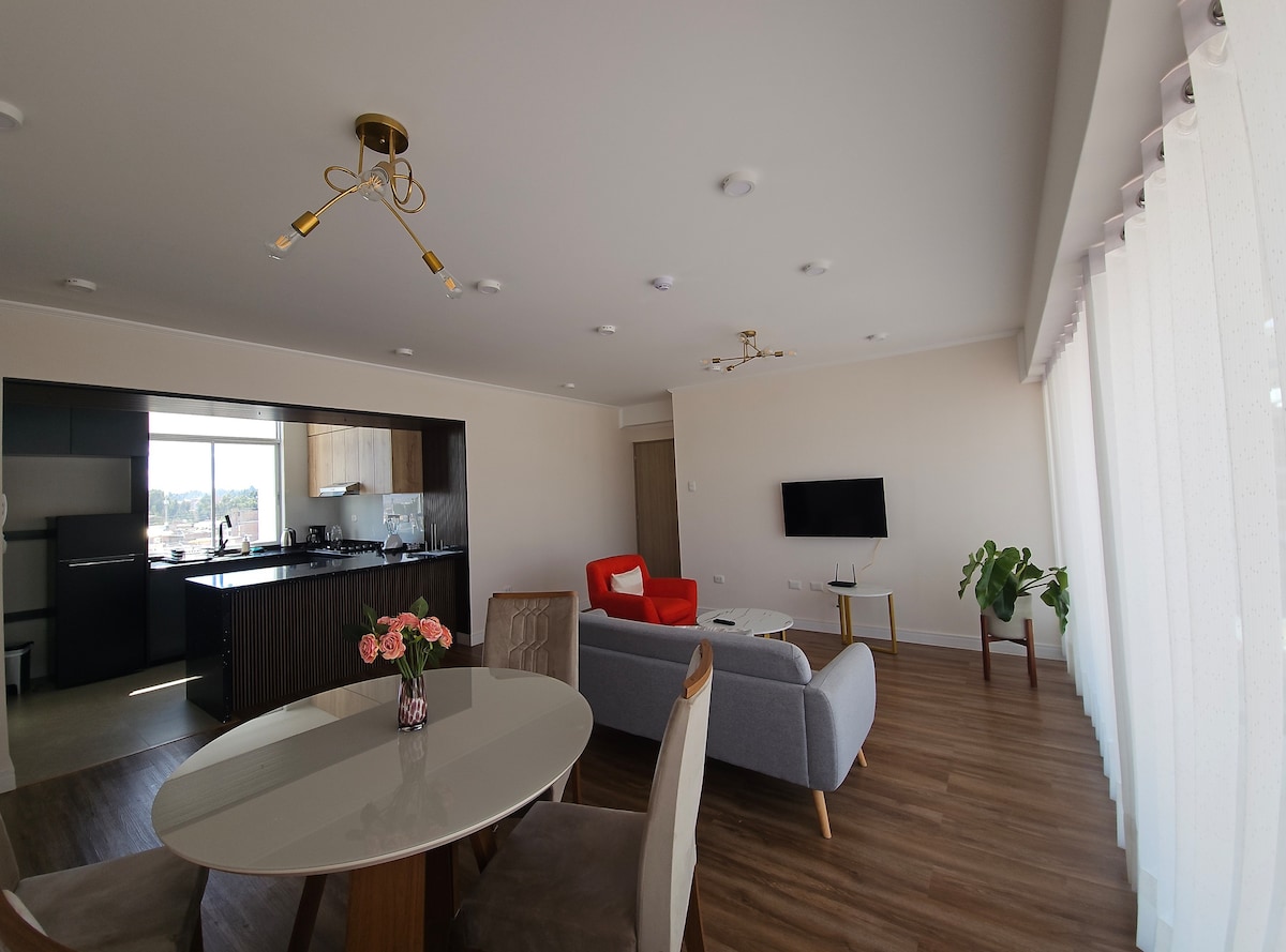 An open living space features a round dining table with five chairs, a gray sectional sofa, and an orange accent chair. A modern kitchen area with dark cabinetry is visible in the background. Large windows allow natural light to brighten the area, complemented by a small potted plant.