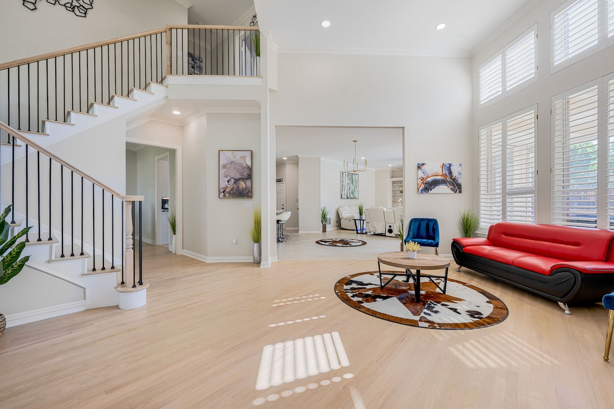 An open and airy living area is highlighted by large windows allowing natural light to flood the space. A modern red sofa contrasts with neutral tones, while a round coffee table sits atop a decorative area rug. A staircase with sleek railings leads to the upper level.