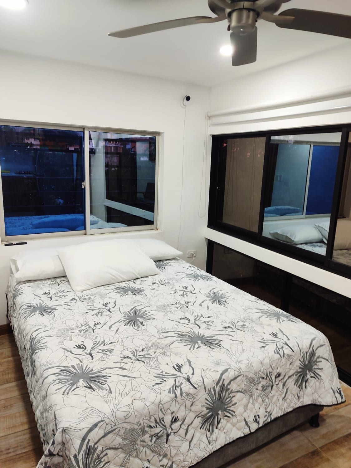 A bright sleeping area features a spacious bed covered with a floral-patterned blanket. Natural light enters through large windows, offering views of outdoor greenery. A modern ceiling fan provides air circulation, while an additional window reflects the interior's clean lines.