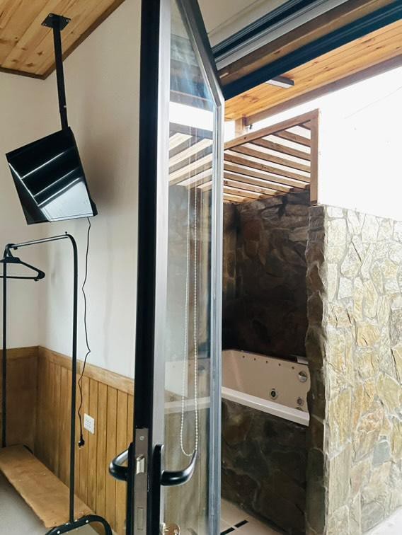 A spacious area is presented with a large glass door leading to an outdoor jacuzzi. Natural stone walls create a rustic ambiance, while a sleek television is mounted in the room. A minimalist bench and a hanging rack are visible, providing a practical design.