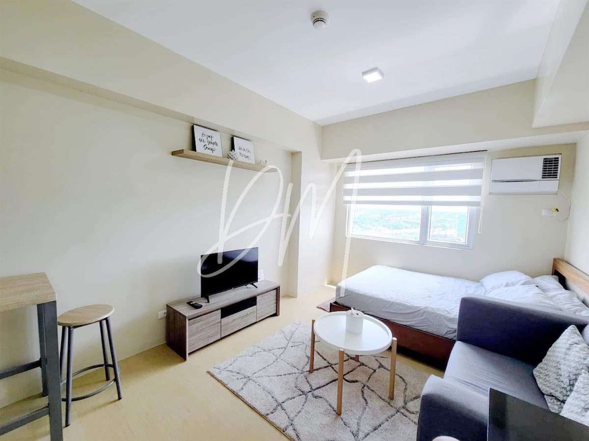 A compact studio apartment is characterized by a neutral color palette. A spacious bed is positioned against the wall, while a small coffee table sits on a patterned rug. An entertainment unit accommodates a television, and large windows allow for ample natural light.