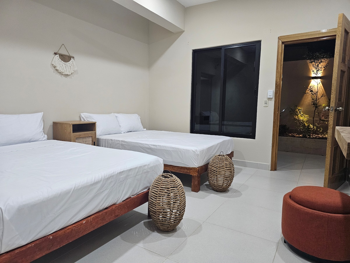 The room features two spacious beds with white linens, positioned to allow easy movement. A natural wood side table stands beside one bed, while two woven baskets add texture to the space. A large window offers a view of a small outdoor area, and a wooden door leads outside.