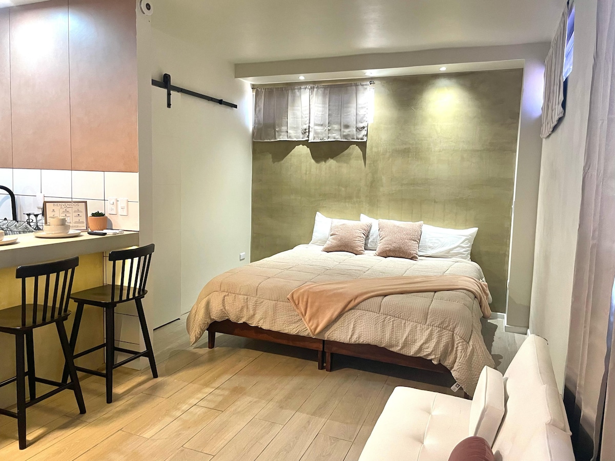 A spacious room is highlighted by a large bed adorned with neutral-colored bedding and several accent pillows. A small dining area with two chairs is visible, alongside a modern couch. Soft lighting is provided by recessed fixtures and natural light coming from a window.
