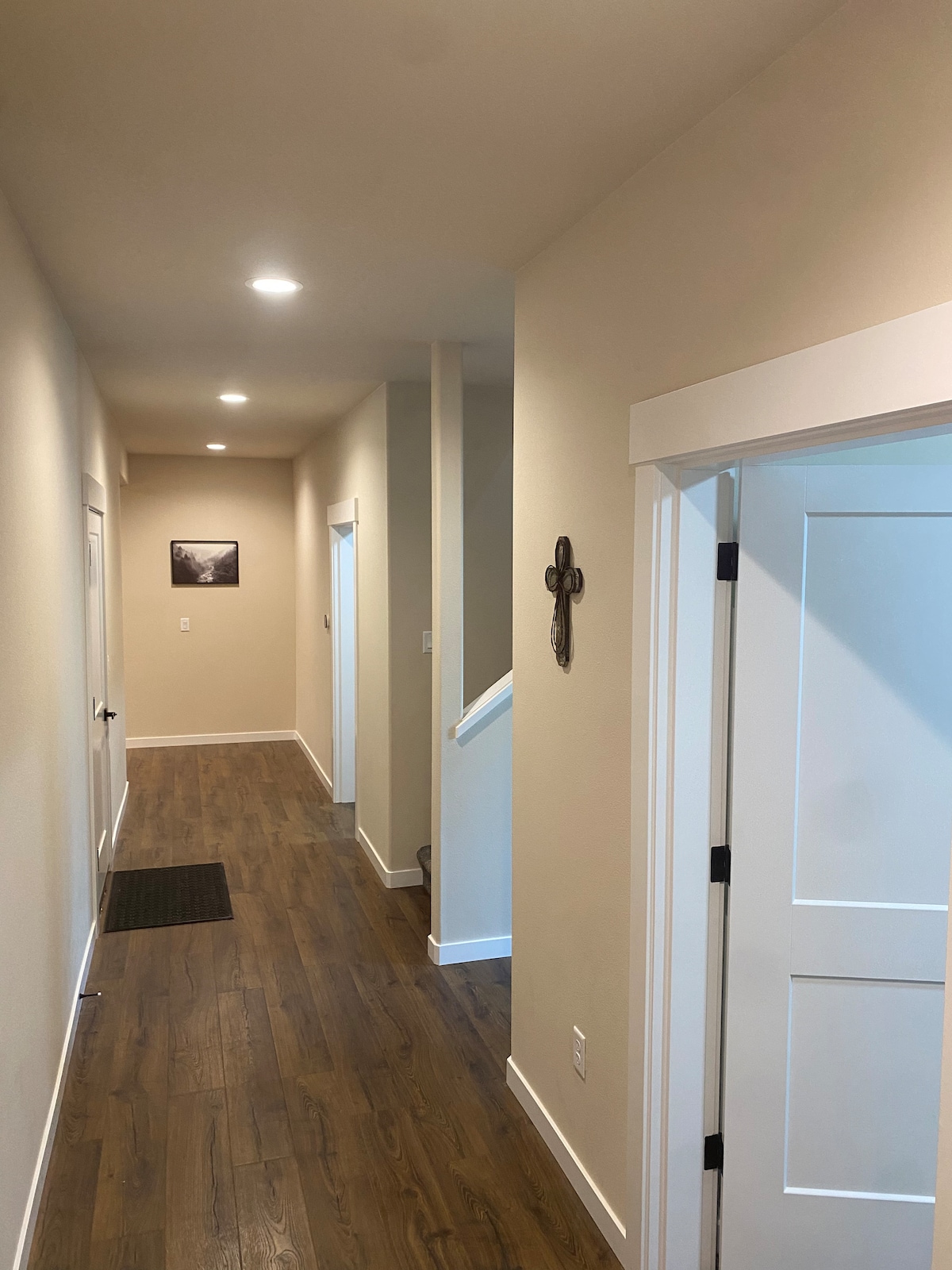 A well-lit hallway features warm wooden flooring and neutral-colored walls. Doorways on either side lead to additional rooms, while a staircase is visible at the end. A decorative wall cross adds a subtle touch of character to the space.