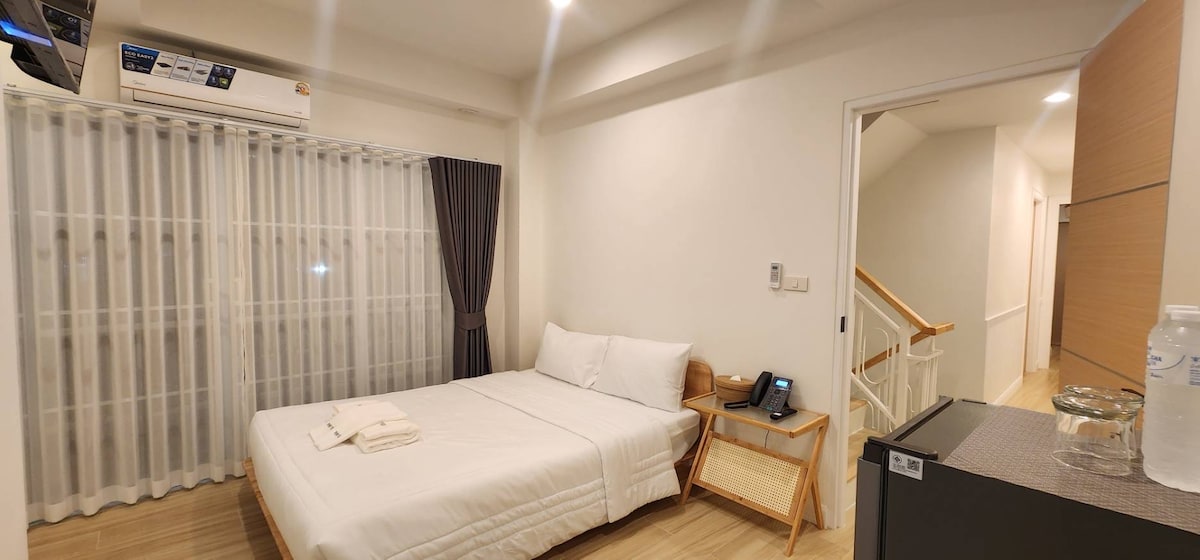 A bright and minimalist bedroom is featured, showcasing a spacious bed neatly made with white linens. Natural light filters through sheer curtains at the window. A small bedside table holds a phone, while a refrigerator stands nearby. The room's layout includes a glimpse of a hallway.