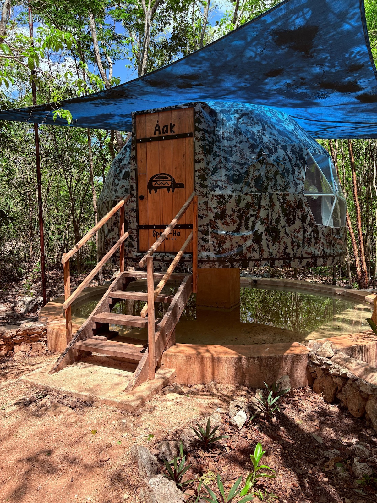A geodesic dome is positioned on a raised platform, surrounded by lush greenery. A wooden stairway leads up to the entrance, where a sign displays the name 'Aak.' A blue canopy offers shade, complementing the natural surroundings.