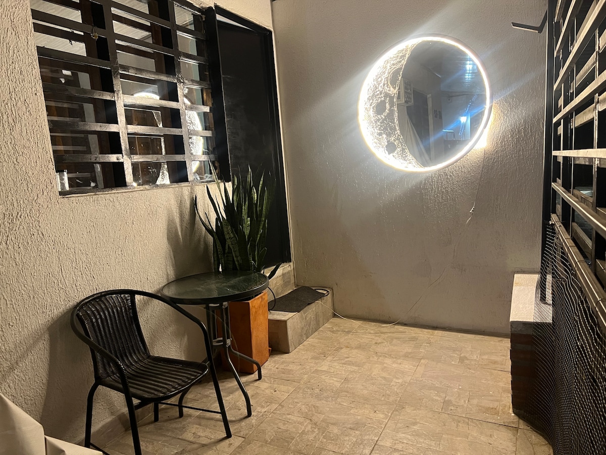 An outdoor seating area features a round table with a glass top and a black chair. A large circular mirror hangs on the wall, reflecting light from the surrounding space. Lush greenery is present, adding an element of nature to the setting.