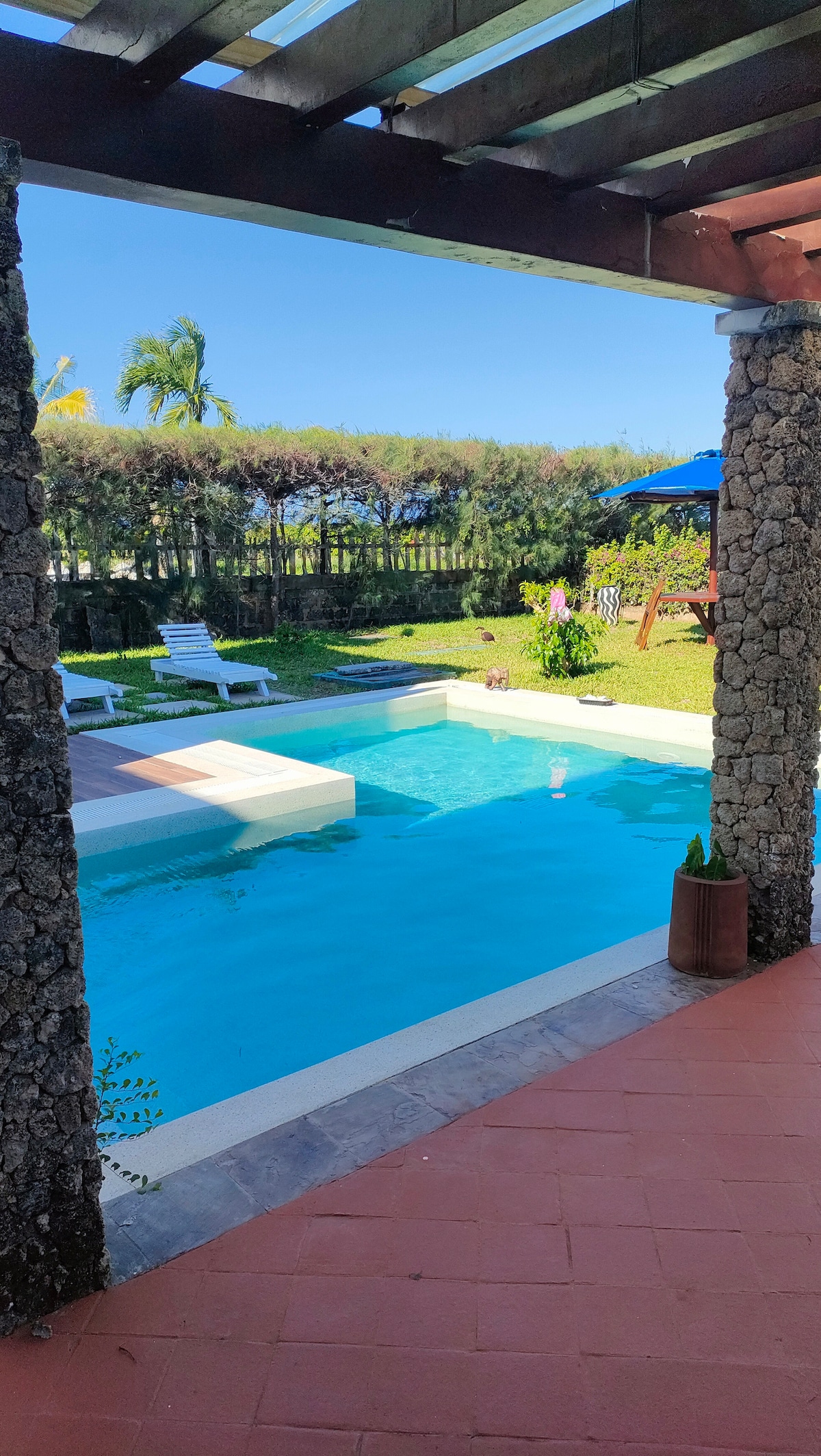 A private pool area is framed by stone pillars and offers a view of the well-maintained garden. Loungers are positioned around the pool, and vibrant greenery provides a natural backdrop. A blue umbrella and a swing set are visible in the distance under clear skies.