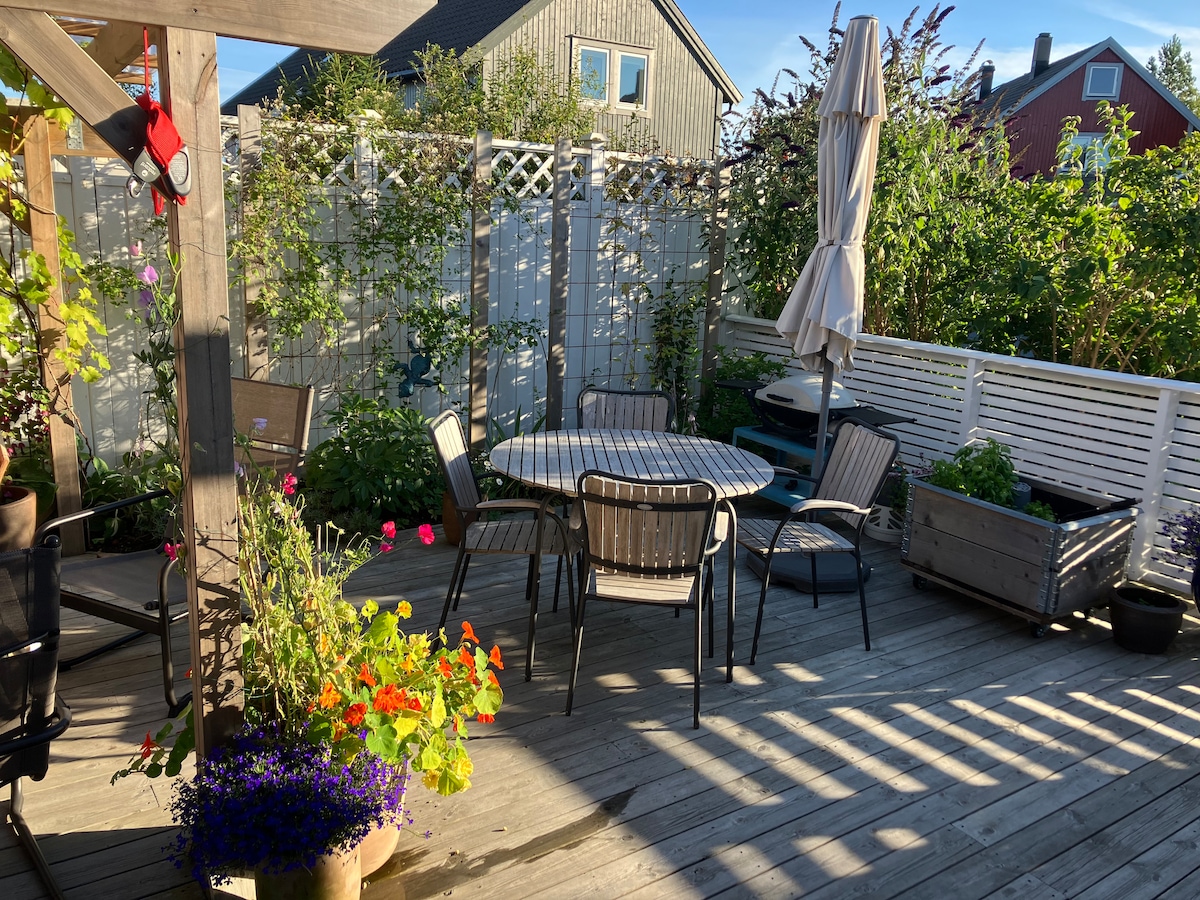 A private outdoor patio is shown, featuring a round table surrounded by several chairs. Potted plants with vibrant flowers add color, while a large umbrella provides shade. Wooden decking creates a warm ambiance, framed by greenery and a trellis.