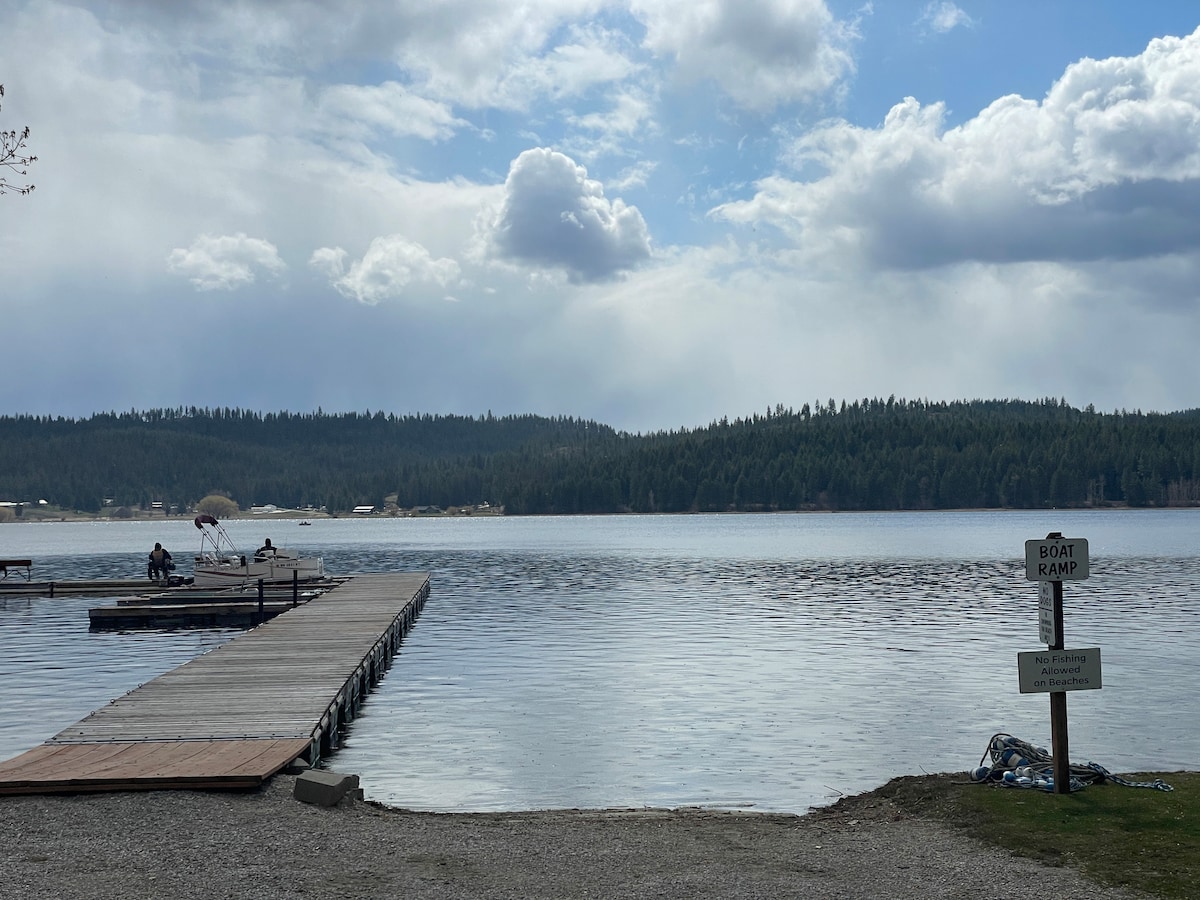 A serene lakeside view captures a wooden dock extending into the calm water, with gentle ripples reflecting the sky. In the distance, a couple is seated on the dock. A boat ramp sign is positioned on the grassy edge, indicating access to the water.