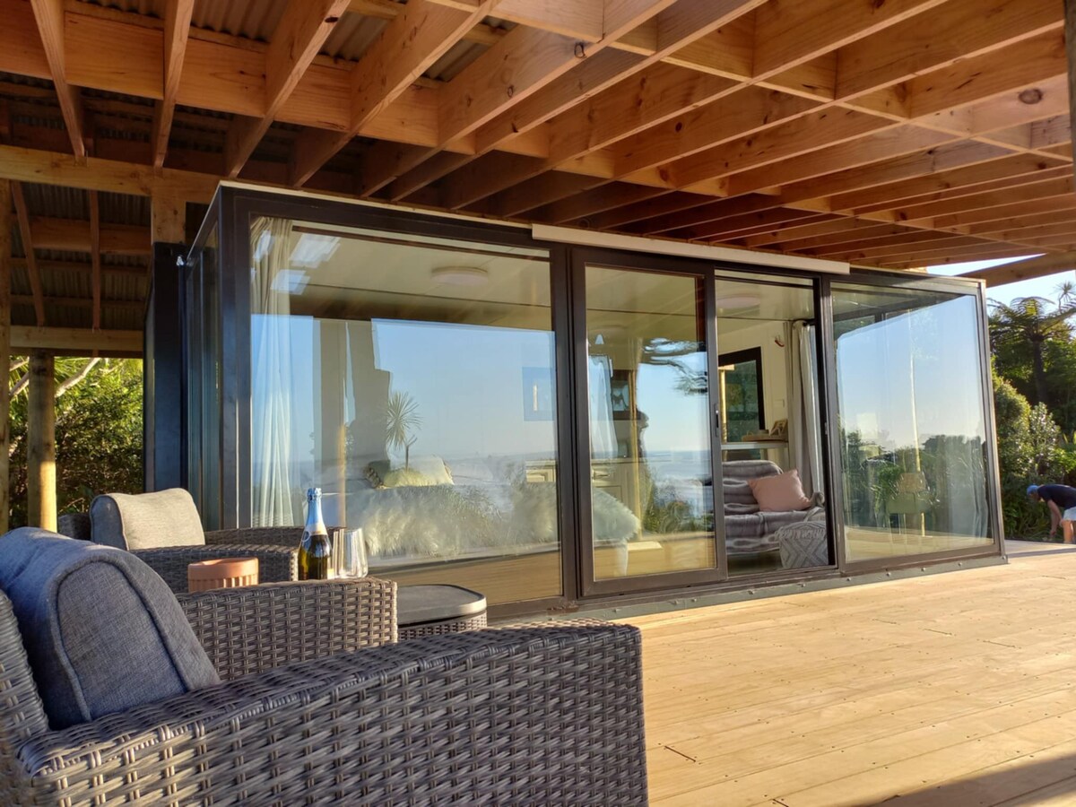 A modern studio is showcased with large glass panels that create a seamless connection to the outdoor deck. Natural light floods the interior, highlighting a cozy seating area furnished with comfortable chairs. A bottle of champagne sits on a small table, suggesting a relaxed atmosphere.