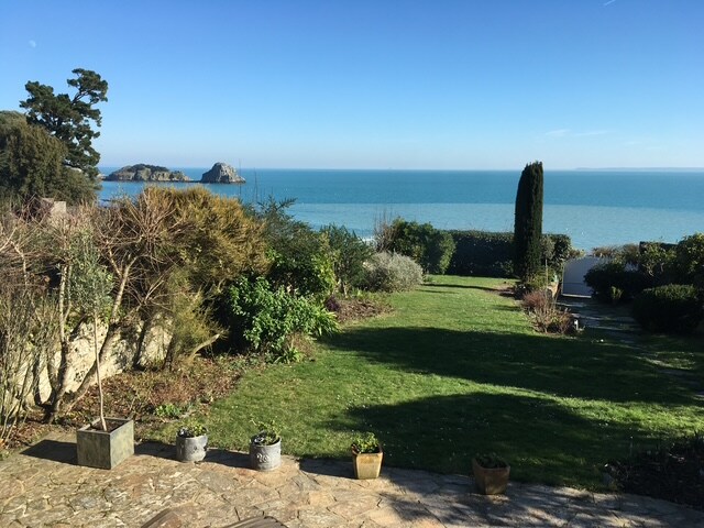 The serene garden landscape is viewed, featuring a well-maintained lawn and assorted greenery. Ocean waves can be seen gently lapping at the shore, with distant rocky formations enhancing the coastal scenery under a clear blue sky.