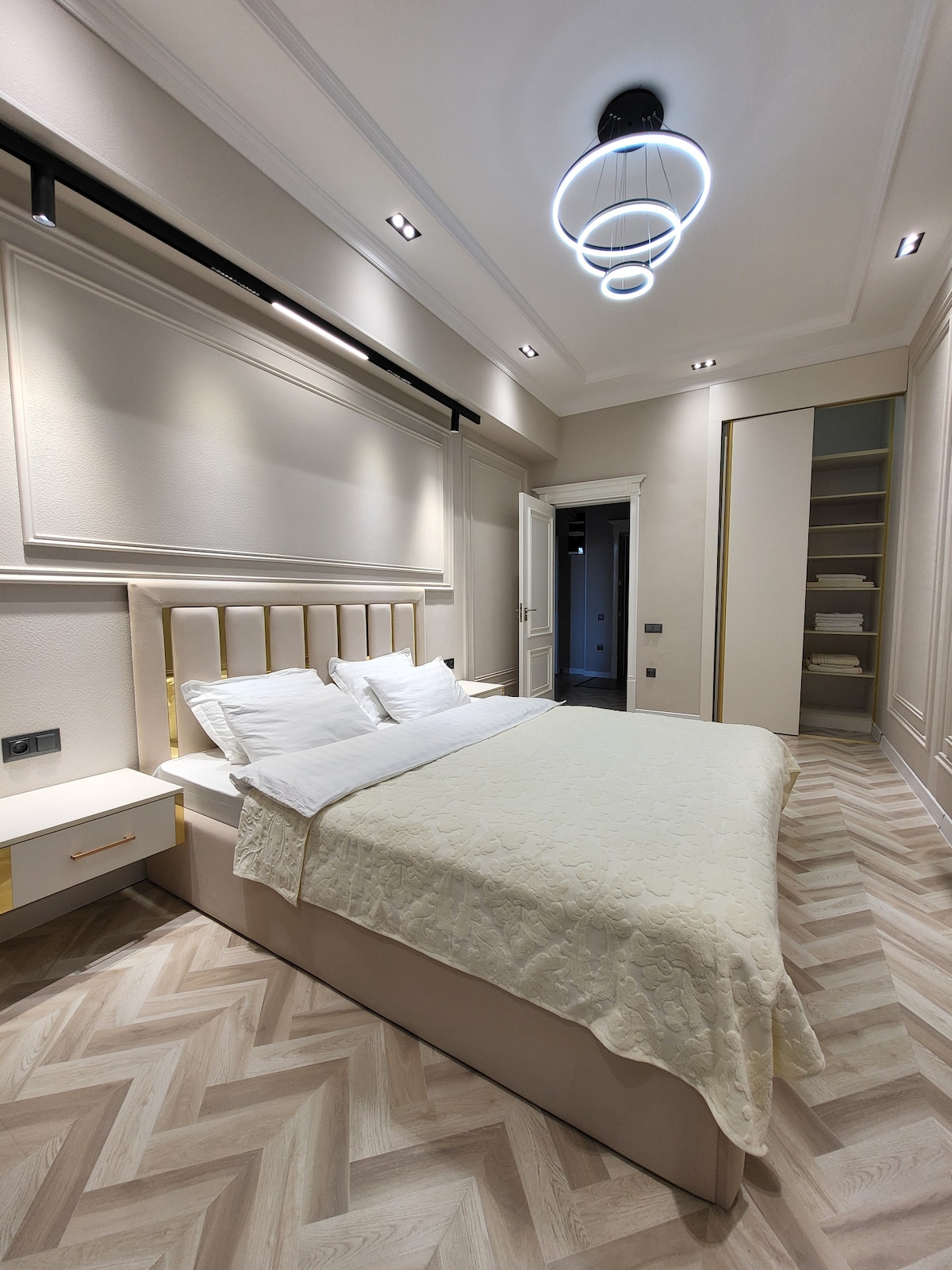 A well-appointed bedroom is shown, featuring a spacious bed with a light-colored coverlet and several pillows. Elegant lighting fixtures are mounted on the ceiling, complementing the neutral wall tones. A sleek nightstand is positioned beside the bed, and a closet is visible in the background.
