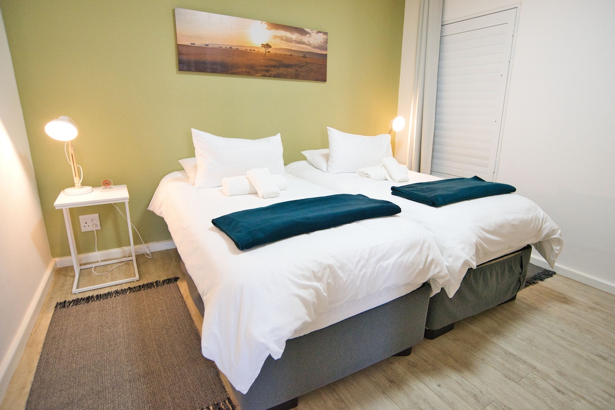 The room features two spacious beds with crisp white linens, set on a light wooden floor. A piece of artwork depicting a sunset hangs above the beds, while small bedside tables with lamps provide soft lighting. A neutral-colored rug adds warmth to the space.
