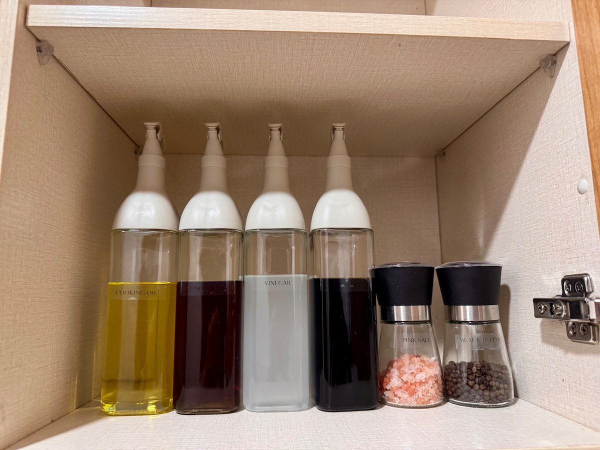 A neatly organized shelf features several clear containers filled with oil, vinegar, and spices. Two grinders are visible on the right, one filled with black pepper and the other with pink salt, providing a variety of cooking essentials.