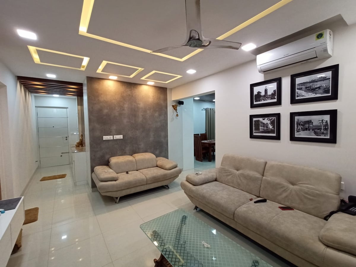 A modern living area features a neutral-colored sofa set arranged around a glass coffee table. The ceiling is highlighted by recessed lighting, creating a warm ambiance. Black and white framed photographs adorn the walls, and an air conditioning unit is mounted above the seating area.
