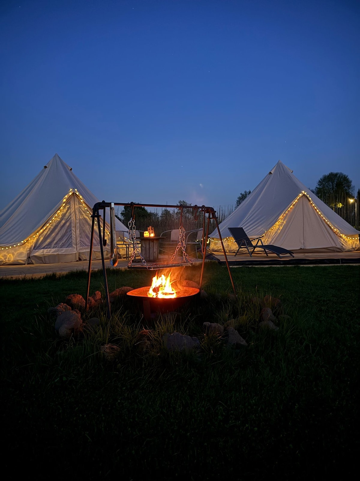 Two illuminated tipi tents are set against a deep blue evening sky. A fire pit with a vibrant flame is positioned in the foreground, surrounded by stones. Soft lights adorn the tents, creating a warm ambiance for outdoor gatherings.