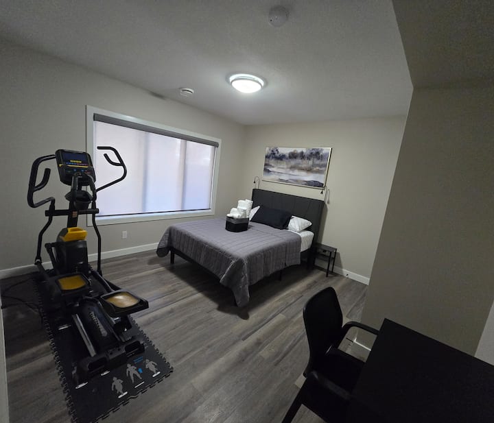 Pogled iz spavaće sobe 1 s ulaznih vrata na kojima se vidi oprema za vježbanje, zidna slika, veliki prozor, radna stanica, krevet, košara s ručnicima, svjetla na noćnom ormariću sa svake strane i noćni ormarić sa svake strane.