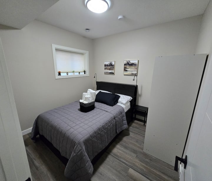 Pogled iz spavaće sobe 2 s ulaznih vrata s ručnicima u košari na krevetu, stalak za krevet s desne strane kreveta, 2 svjetla na stalku sa svake strane kreveta, dekoracija za slike, stojeći ormar i prozor.