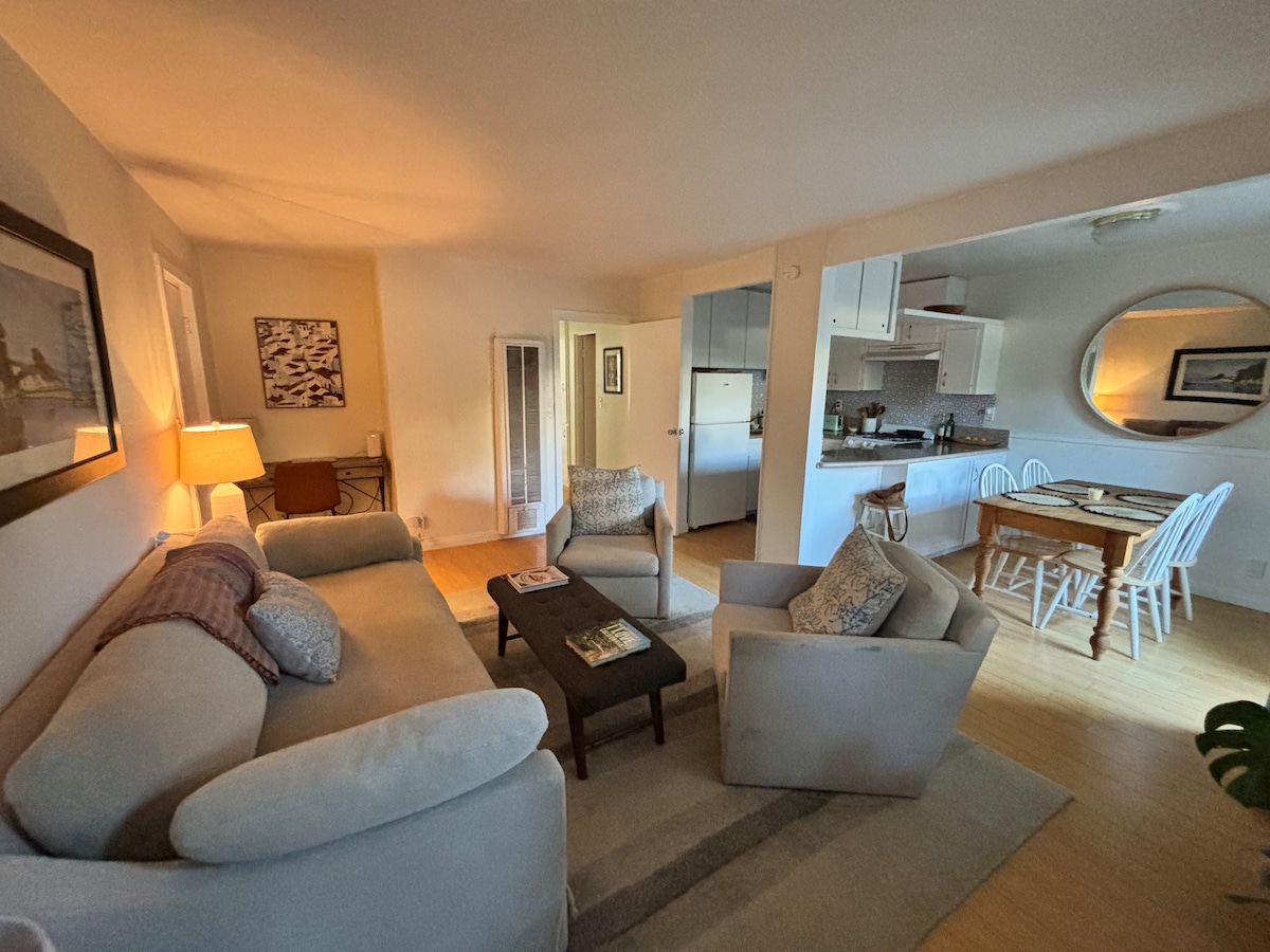 A welcoming living area is presented with a light-colored sofa and two armchairs, arranged around a coffee table. A dining area with a round table and white chairs is visible in the background, along with a well-equipped kitchen. Soft lighting emanates from a nearby lamp.