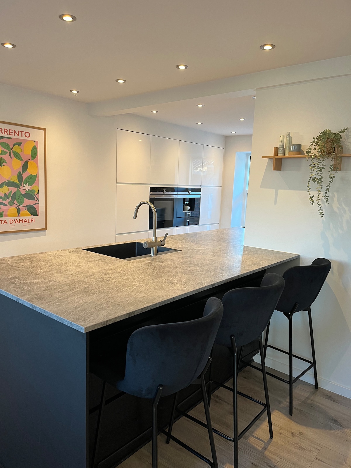 A sleek kitchen features a modern island with a textured countertop and four black bar stools. Bright overhead lighting illuminates the space, while minimalist cabinetry lines the walls, complementing the clean design. A small shelf holds decorative items and greenery, adding a touch of color.