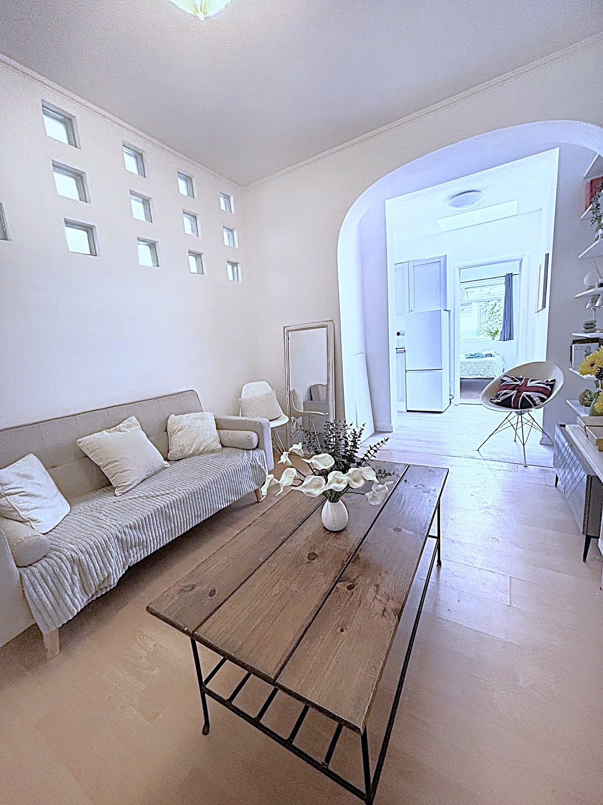 A lounge area features a light-colored sofa with cushions, accompanied by a wooden coffee table at its center. A mirror and decor items adorn the space, with natural light filtering through nearby windows. An archway leads to the kitchen area visible beyond.