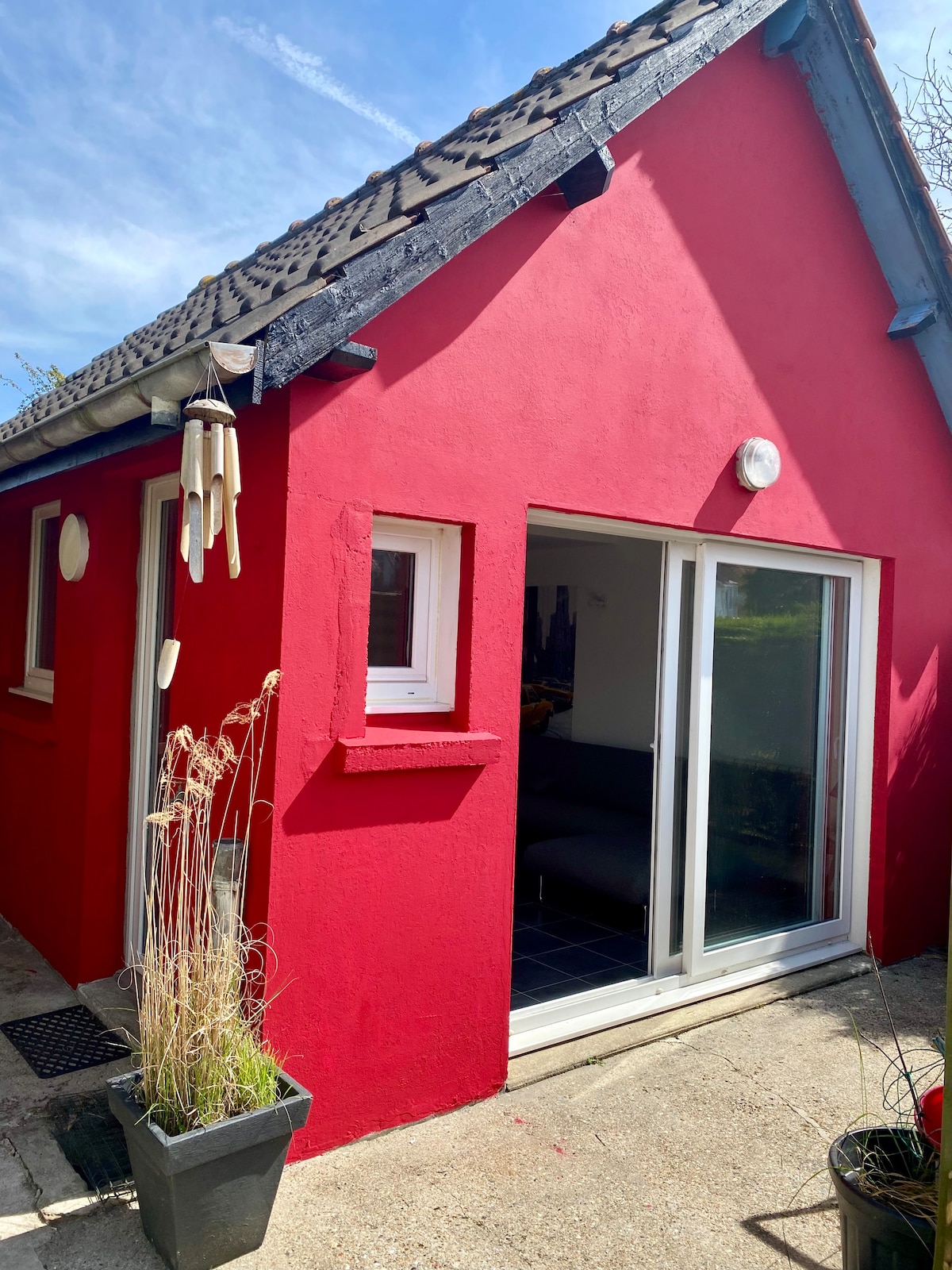 A small red house is featured, showcasing a sloped roof and two windows on the facade. The entrance includes sliding glass doors that lead to the living space. Potted plants with dried grasses are positioned at the entrance, adding a natural element to the vibrant exterior.