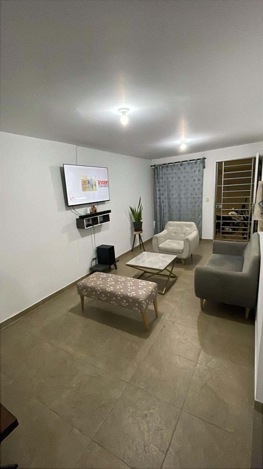 A modern living area is showcased with two light-grey armchairs and a white ottoman with a floral pattern. A flat-screen TV is mounted on the wall, and a rectangular glass coffee table stands in the center. Natural light filters through a partially draped window adorned with light curtains.