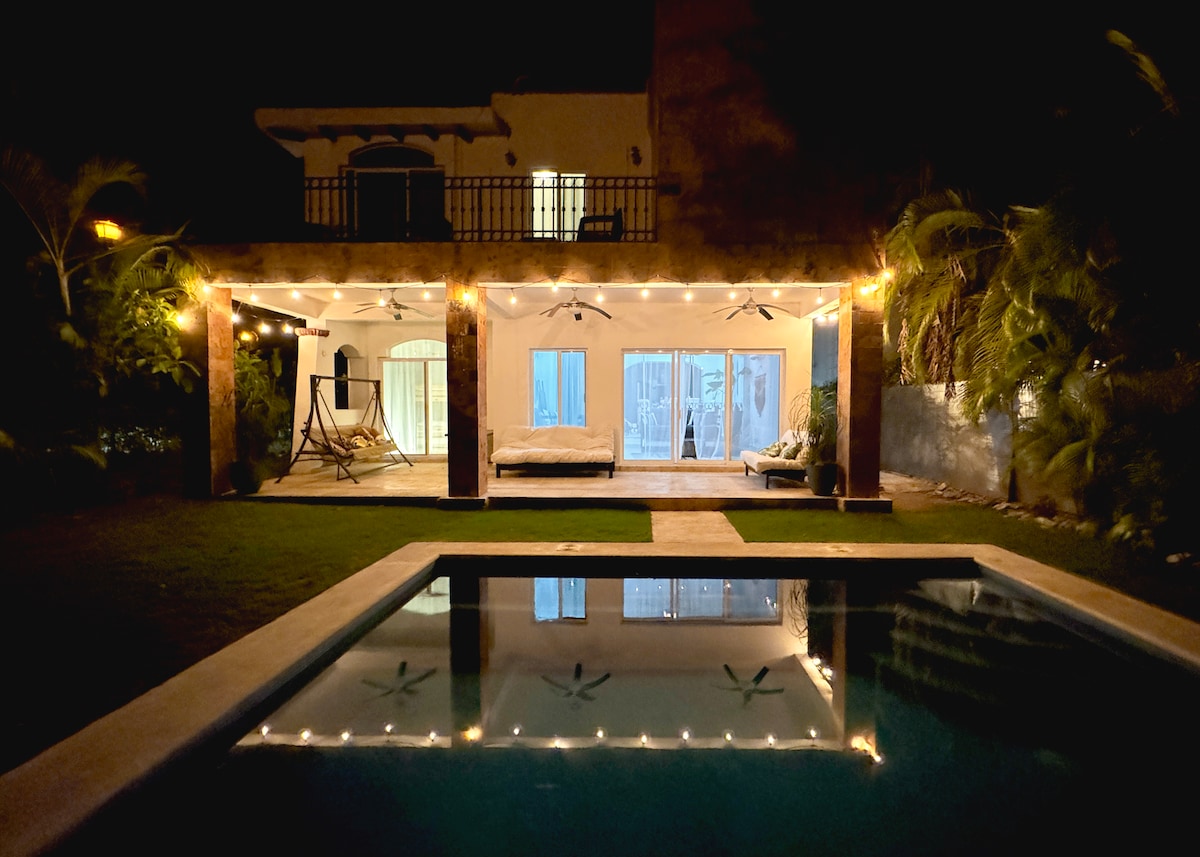 The villa is illuminated at night, showcasing a spacious outdoor area with a private pool and tasteful lighting. The covered patio features a comfortable seating arrangement and a hammock, while large glass doors provide a view of the interior space, blending indoor and outdoor living.