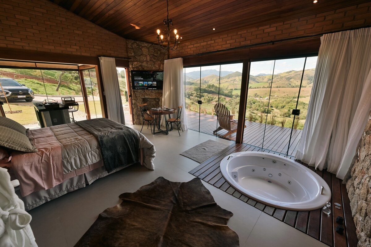 A spacious room features a queen-size bed adorned with luxurious bedding and a plush blanket. Large glass doors provide panoramic views of the surrounding mountains and open to a deck. A circular hot tub is positioned nearby, complementing the rustic stone and wood accents throughout the space.