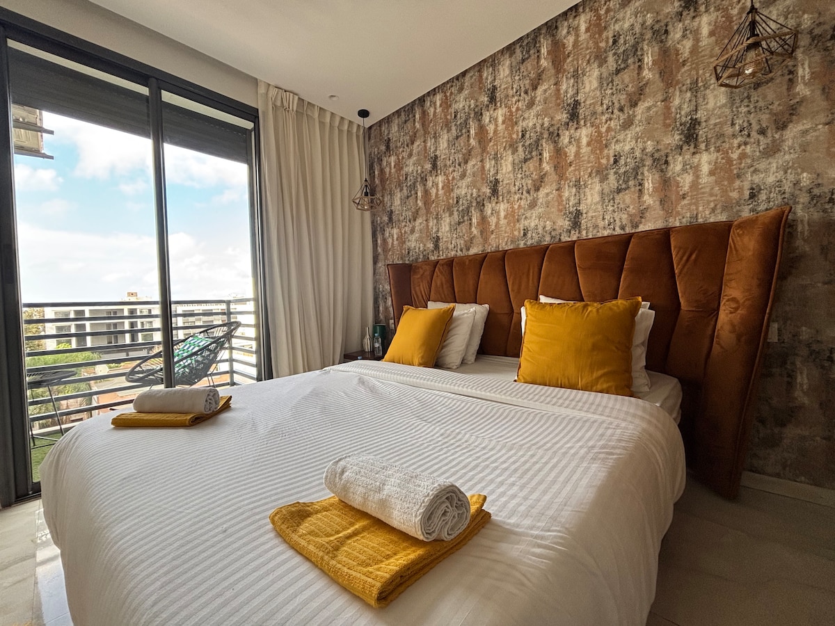 A spacious bedroom features a large, upholstered bed adorned with yellow accent pillows. Soft linens are neatly arranged, and two rolled towels are placed on the bed. The room is brightened by natural light coming through large glass doors that open to a balcony.