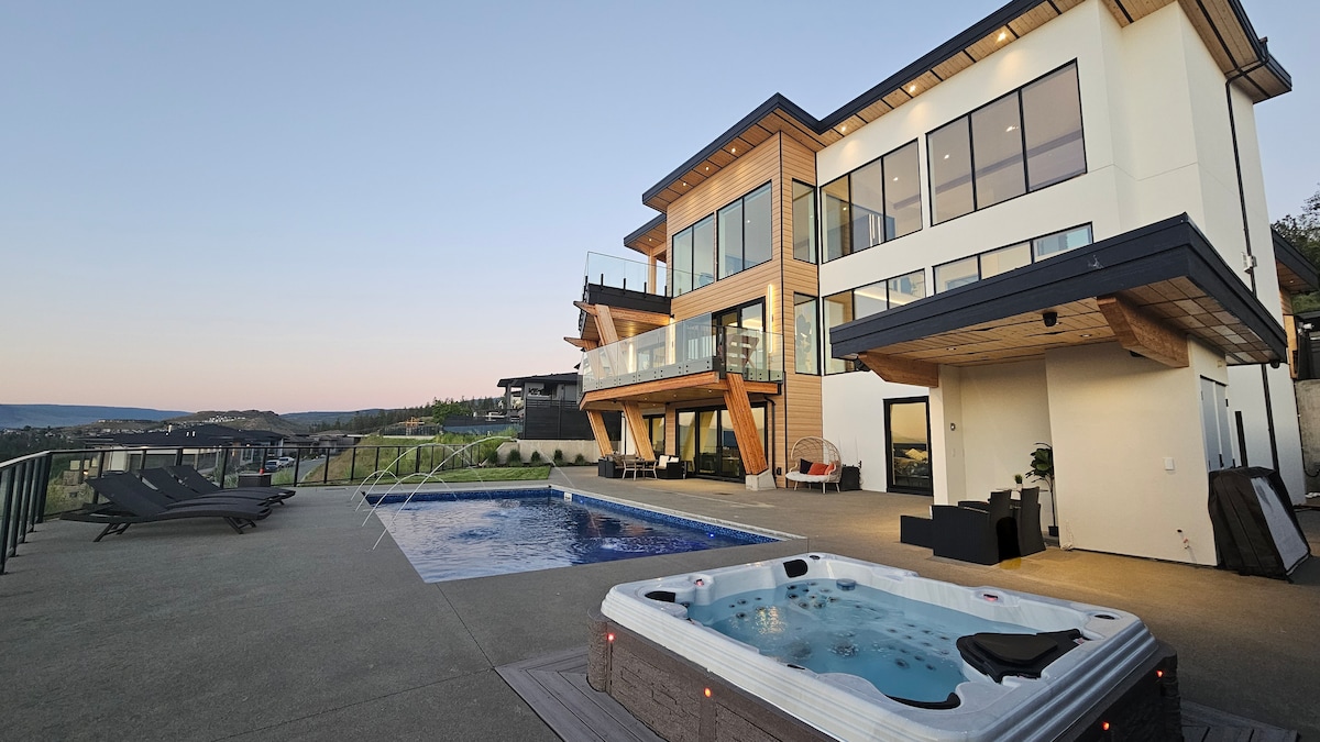 An expansive outdoor area is presented, featuring a large saltwater pool alongside a hot tub. The modern home showcases multiple levels with floor-to-ceiling windows. The patio is furnished with lounge chairs, providing a space for relaxation. Scenic views stretch across the landscape.