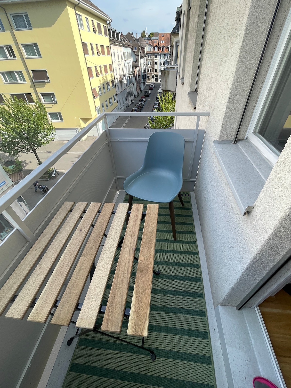 A small balcony features a wooden slatted table and a single light blue chair. A green-striped rug adds comfort underfoot. The view outside includes a glimpse of the neighborhood, with buildings lining the street below.