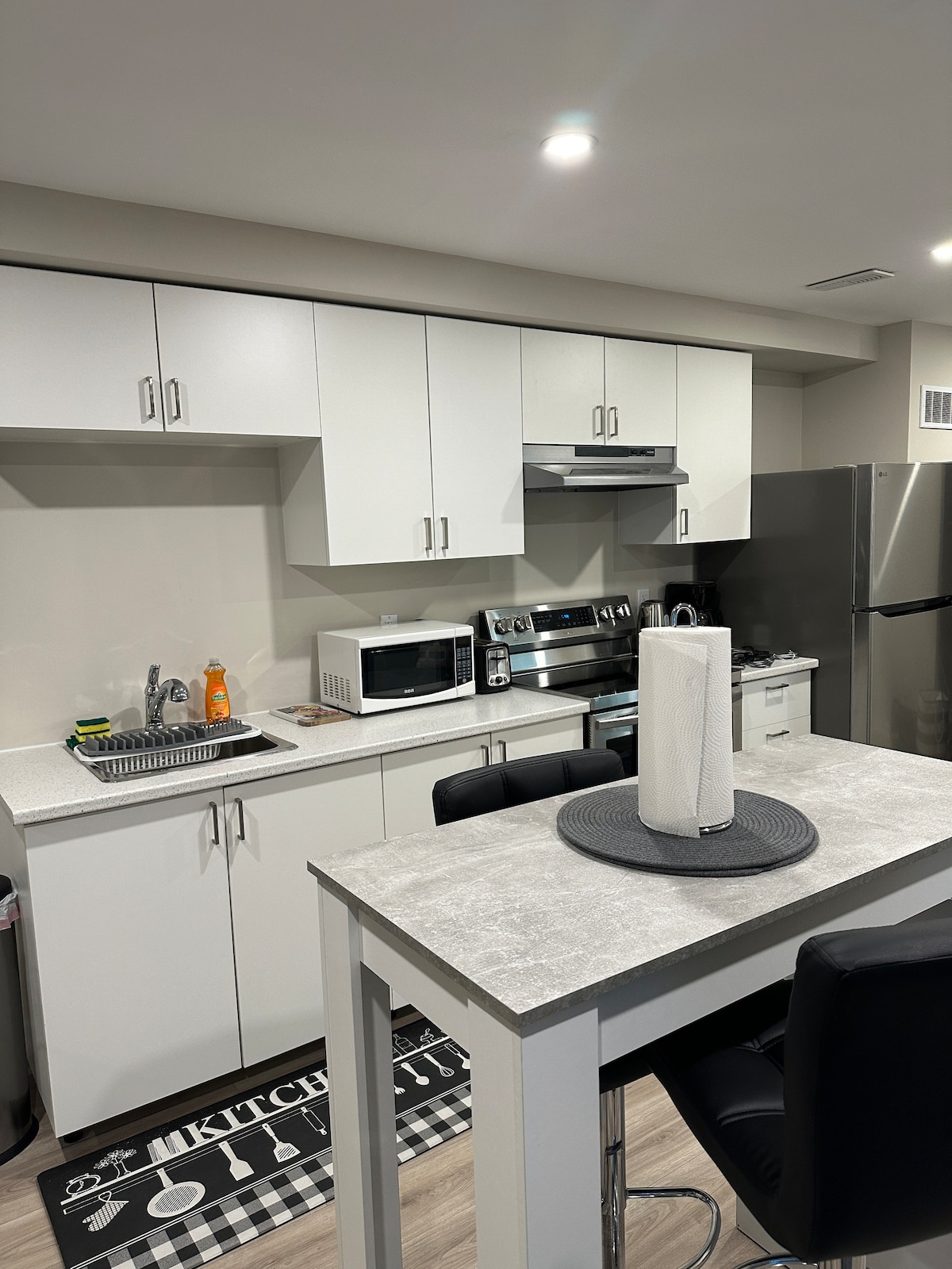 A modern kitchenette equipped with a stainless steel refrigerator, microwave, and oven is set against sleek white cabinetry. A small dining table is highlighted in the center, featuring a decorative placemat and black chairs. Ample counter space and organized storage contribute to a tidy atmosphere.