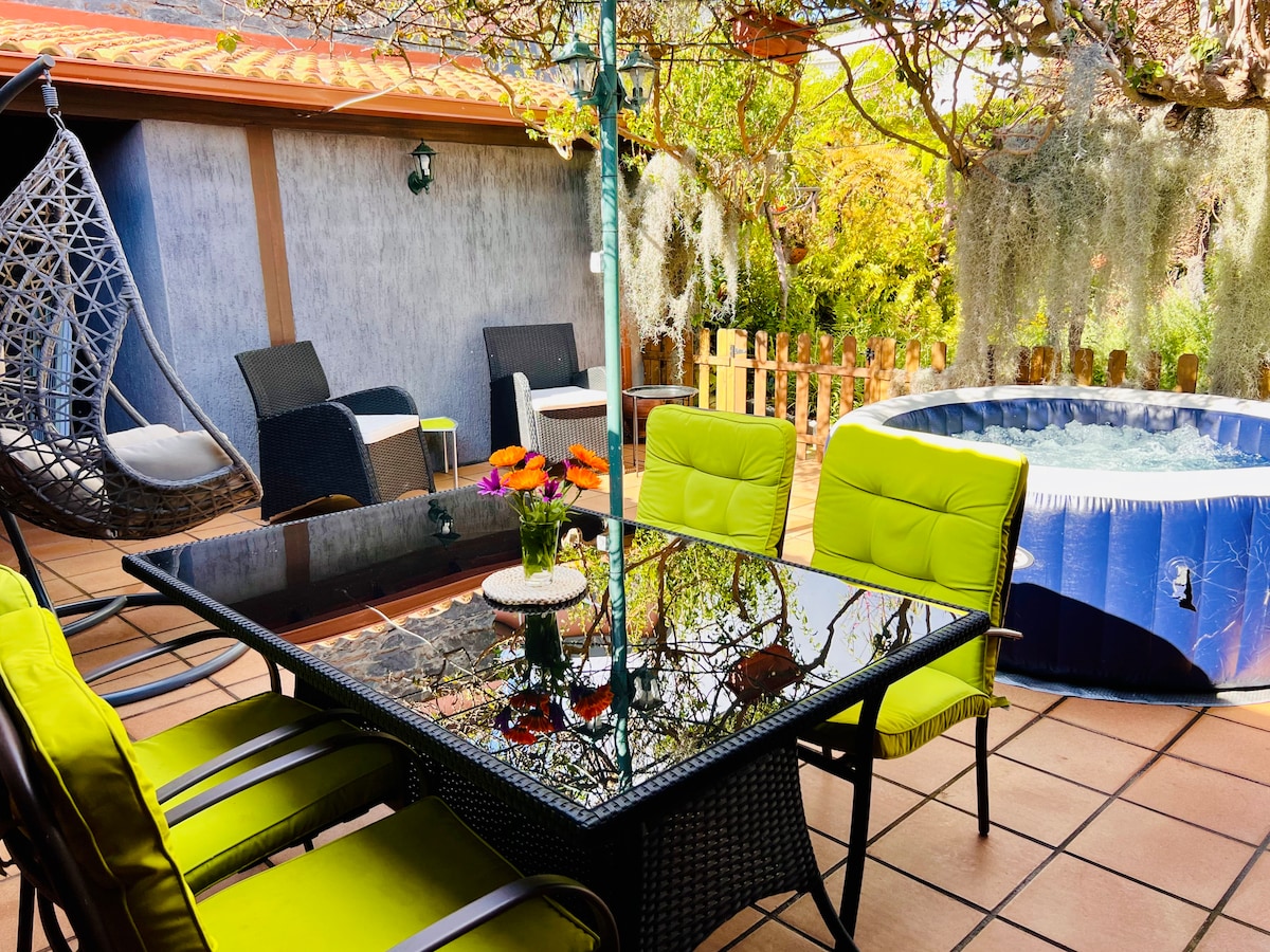 An outdoor patio space is presented, featuring a large glass table surrounded by four bright green chairs. A hot tub is visible to the right, and a hammock swings gently in the corner. Lush greenery and hanging plants add to the tranquil atmosphere.