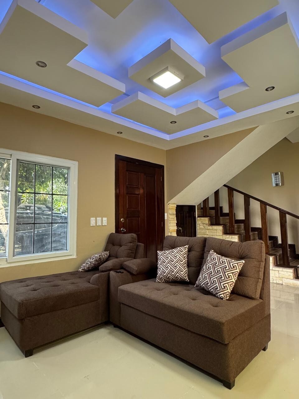 A cozy living area features dark brown sectional sofas adorned with patterned cushions. The room is illuminated by modern recessed lighting and has a large window overlooking greenery. A wooden staircase leads to a second level, enhancing the sense of space.