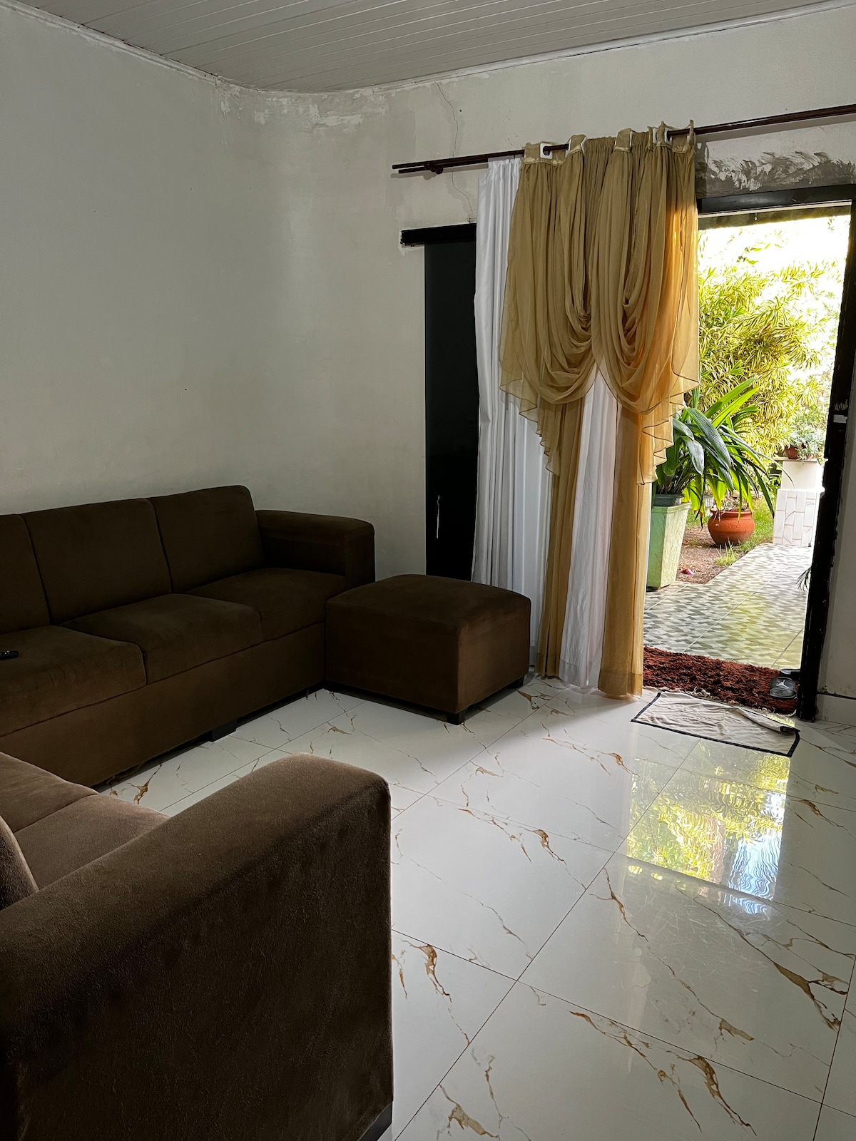 A living room area features a brown upholstered sofa set with a matching ottoman. Natural light enters through a doorway adorned with sheer curtains, revealing an outdoor space with potted plants. The tiled floor reflects the light, enhancing the inviting environment.