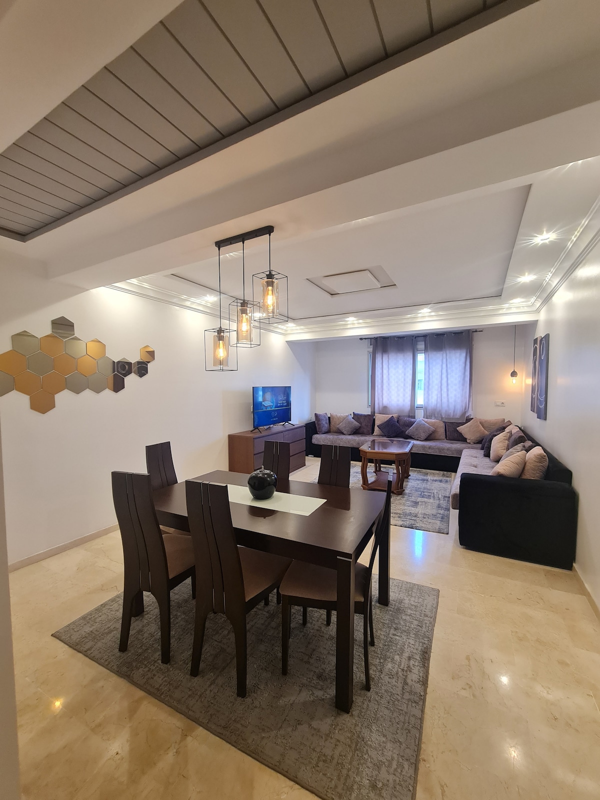 A dining area featuring a dark wood table with six chairs is visible. Soft lighting is provided by pendant fixtures above. A comfortable seating arrangement with pillows is seen in the corner. Natural light enters through a window, enhancing the inviting atmosphere of the space.